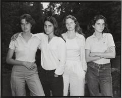 Group portrait of four women standing outdoors. The left two women have their arms wrapped around each other's shoulder. The right woman stands with her arms crossed.