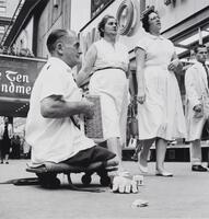 A legless man seated on a dolly on the sidewalk. Two women and a boy are walking by, not noticing him all wearing white.