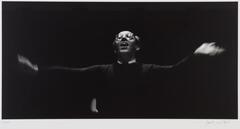 Black background, man with glasses on, hands outstretched conducting an orchestra.