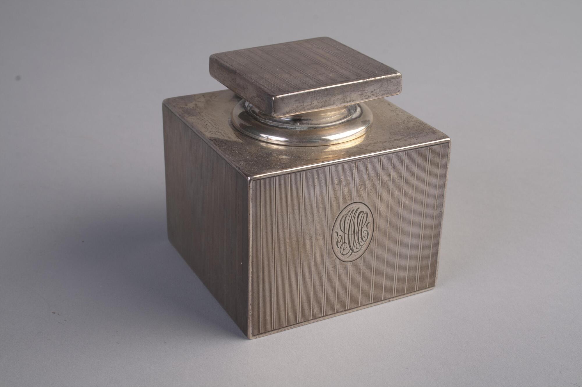 A square, sterling silver inkwell. The base is monogrammed and has etched lines. The lid is flat, square, and also has etched lines.
