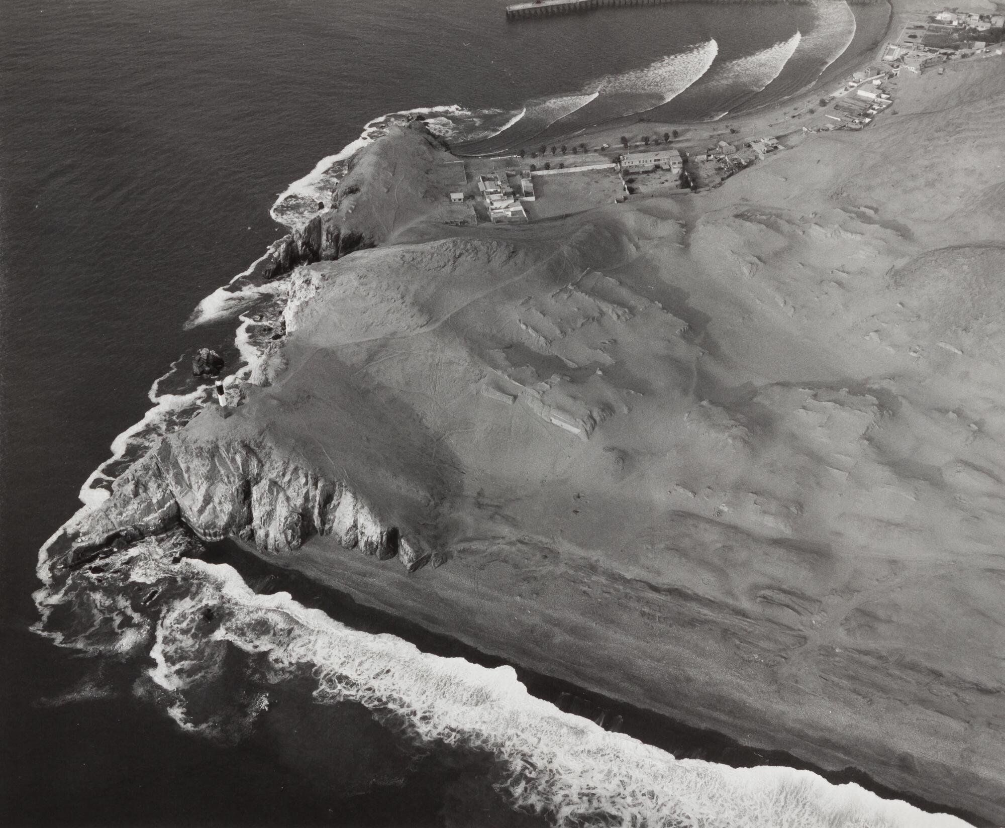 This photograph depicts an aerial view of a coastline on which sits a modern town and the ancient ruins of a city.