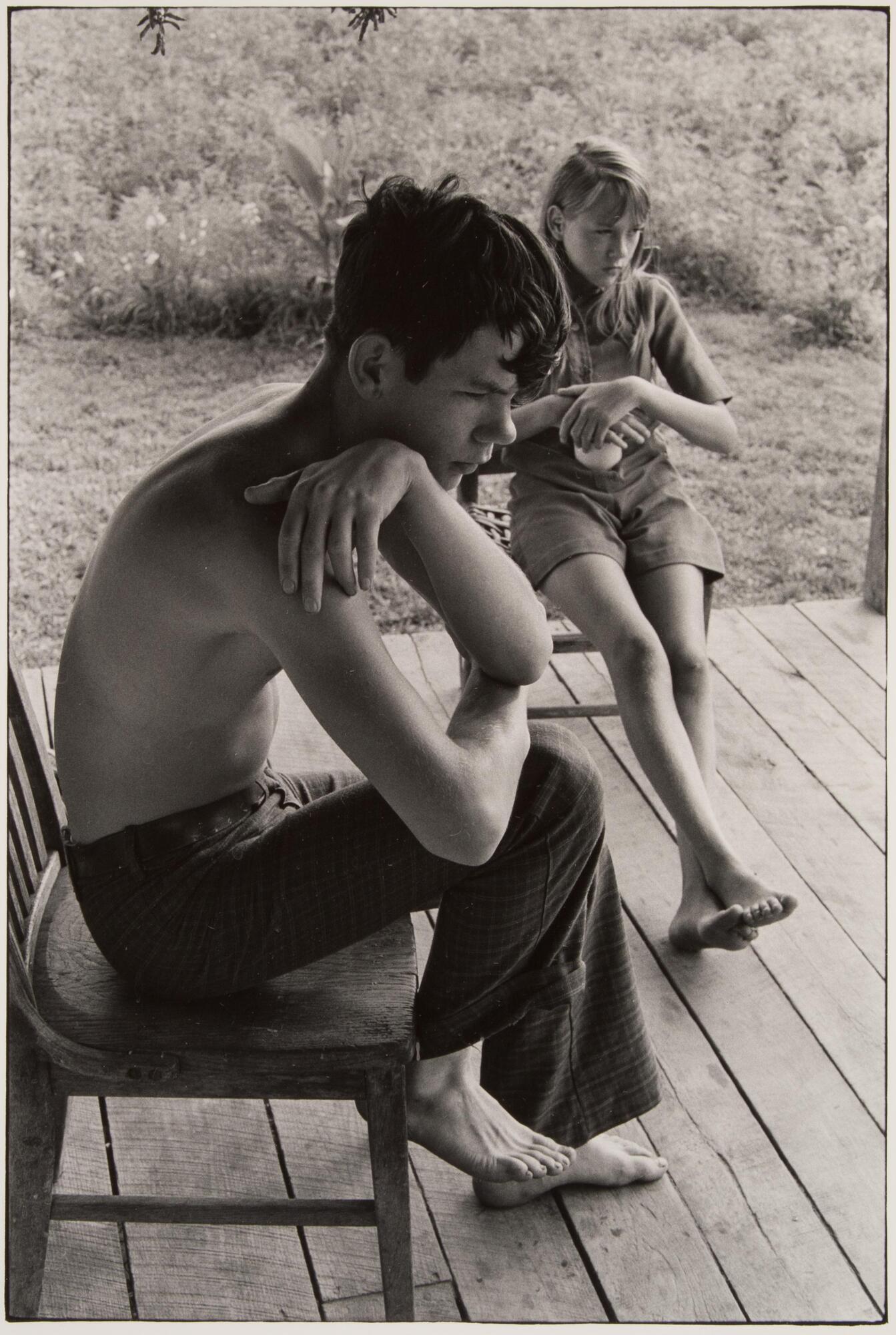 A boy and girl seated on wooden chairs on a porch The boy is shirtless and hunched over, the girl is barefoot.