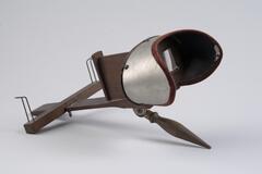 It is a stereoscope viewer made of wood and brass. The viewer part is supported by a wooden handle and has two glass pieces for the viewer to look through. There are two pieces of wood is a cross shape that are also attached to the viewer, the horizontal piece can be slid along the virtical piece and there are two brass supports that hold the stereoscope card.