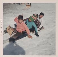 A group of four boys sitting in a line on a sled in the snow. The boy in front wears a pink sweatshirt and the second boy wears a blue shirt and green winter hat. The photographer&#39;s shadow is visible in the lower left corner.