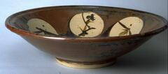 Shallow bowl with unglazed foot. Body of the bowl is decorated with a brown glaze, with circular area on interior of bowl areas where wax resist was applied and tan shows through. In these circles are painted brown stylized floral designs.