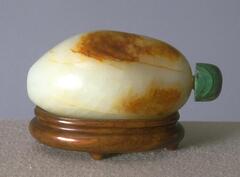An orange and white oval nephrite jade snuff bottle, lying on its side. It has a green stopper and is lying on a wooden stand.
