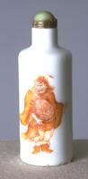 A porcelain white cylindrical shaped snuff bottle. Painted on the front is a design of a red and orange Demon Queller. On the top is a ivory or horn stopper with a brass collar.