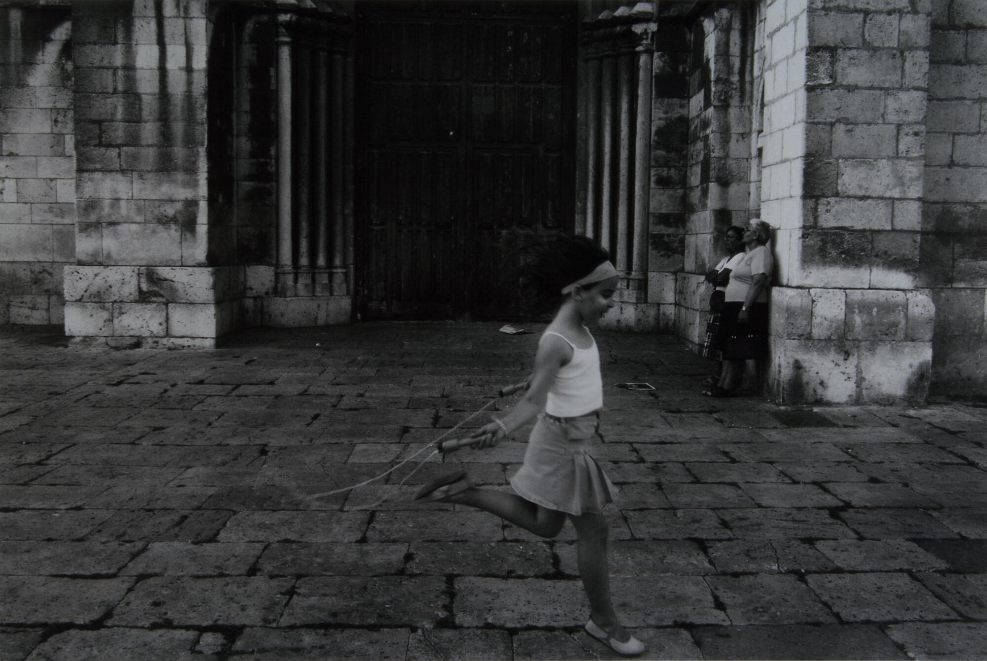 A little girl jumping rope. Two women in the background are leaning up against a building.
