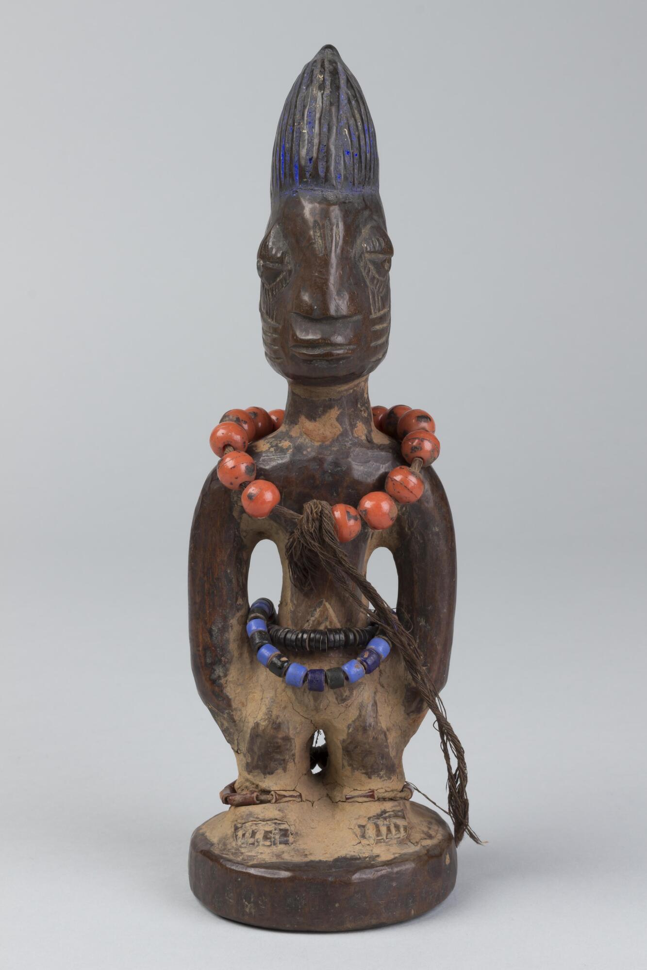 Standing male figure on a round base with hands at the sides. Around the waist are two strings of black and blue beads, a  string of round, orange beads around the neck, as well as a geometric pattern on the abdomen and brass around the ankles. On each cheek are four horizontal grooves topped by three vertical grooves and three vertical grooves. The pupils are metal and the hair is in a conical shape with a small knob and coated in blue pigment. 
