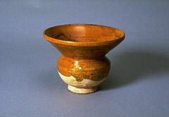 This stoneware spittoon has a globular body on a straight footring, a narrow neck, and tall flaring rim. Three quarters of the body is covered in an amber glaze. 