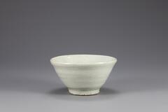 Small porcelain bowl with thick vessel walls, somewhat coarse exterior, and bluish glaze.<br />
<br />
Produced in a regional private kiln in the late 19th century, this white porcelain bowl is has a bright-colored clay body and a wide foot. Sand spur marks are left on the rim of the foot as well as on the inner base, because it was stacked among other bowls, separated by sand supports, in the kiln. It is coated with white opaque glaze and features many pinholes on the outer base due to crawling of glaze.<br />
[Korean Collection, University of Michigan Museum of Art (2014) p.163]