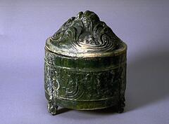 This cylindrical, red earthenware vessel rests on three zoomorphic feet. It is molded in low relief with hills and running animals. Opposing monster masks hold rings as faux handles. The vessel has a high, pointed dome cover moulded with animals running though mountains and clouds. It is covered in a lead green glaze with iridescence and calcification.