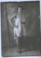 A studio portrait of a young African American woman in 1920s apparel, including a short hairstyle, stockings, necklace, and dress. She stands with her left leg resting on a bench with her right arm akimbo.