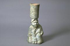 A red earthenware candlestick holder in the form of a large, androgynous human figure, presumably female, kneeling, with a child. There is a cylindrical holder for a candle on the larger figure's head. The candlestick is covered in a green lead glaze, with iridescence and calcification.