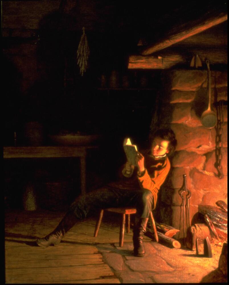 A figure wearing dark boots, dark pants, and brown overshirt reads, sitting on stool with his back to lit fireplace. Set in darkened domestic interior, table and drying herbs visible at back, wooden floorboards and stone of fireplace are illuminated. (Larson 2/5/18)&nbsp;<br />
&nbsp;