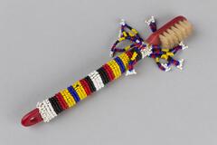 Beaded red toothbrush with striped beading covering in white, black, red, yellow and blue beads. Tassles near the toothbrush head. White string tied through hole at bottom of toothbrush.