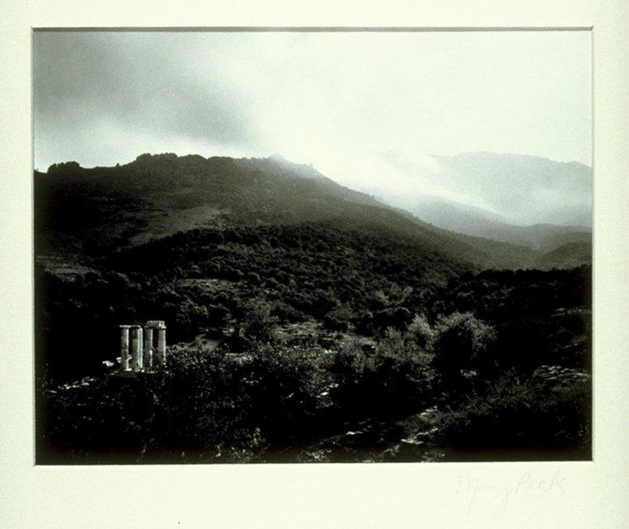 This photograph depicts a temple in the lower left of rolling hills set in a rocky landscape. Clouds sweep over the mountains in the background.
