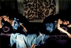 This is a color photograph of two models reclining on a purple satin sofa, a patterned backdrop behind them. They each wear blue satin night gowns. The composition is symmetrical, with one woman on each end of the sofa, leaning on the cushions and looking out of frame.
