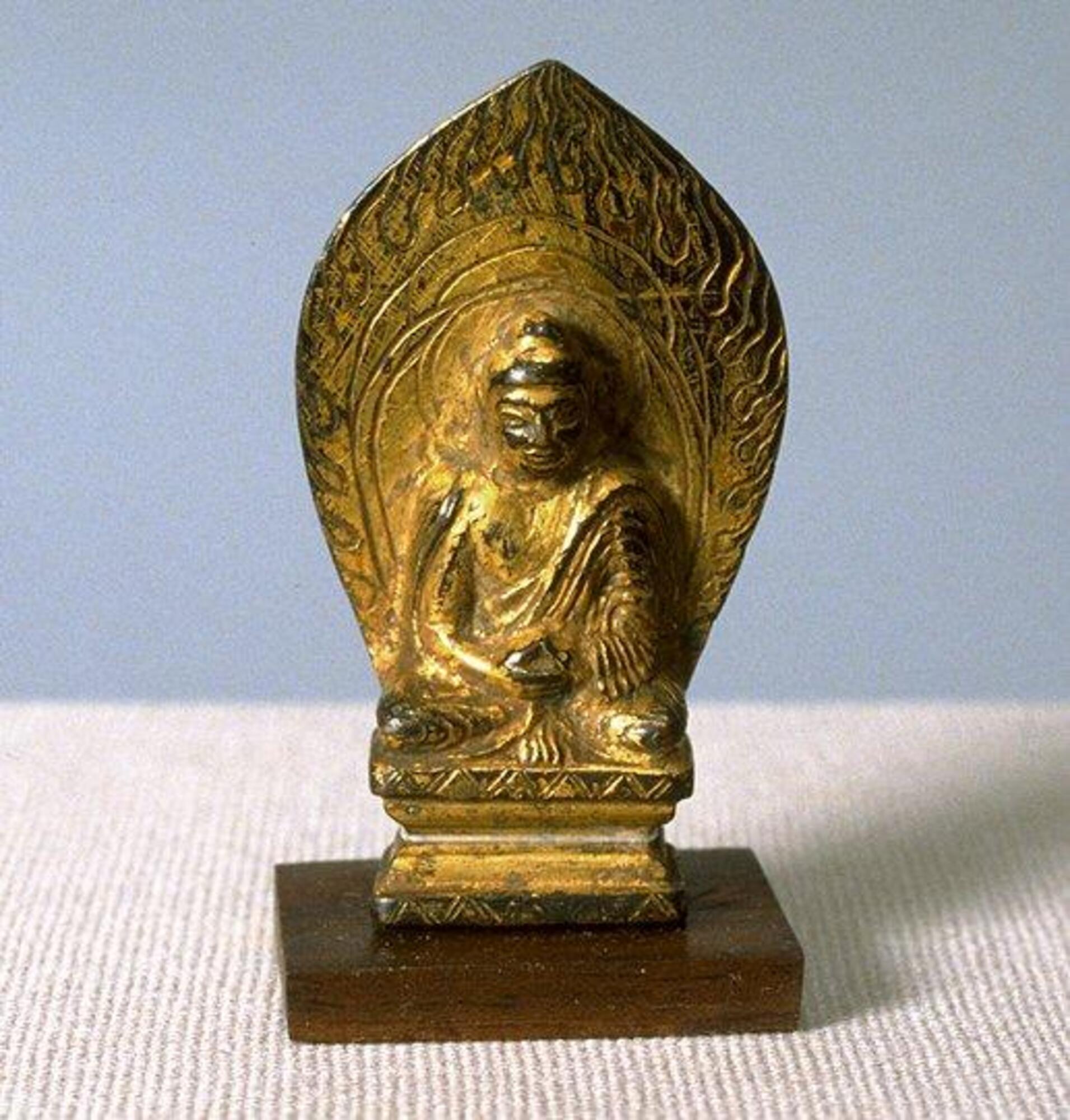 Small gilt bronze seated Buddha with Indian and Central Asian characteristics, including the pedestal he is seated on, folds of his robe, and the incised flames in the body halo encompassing him.