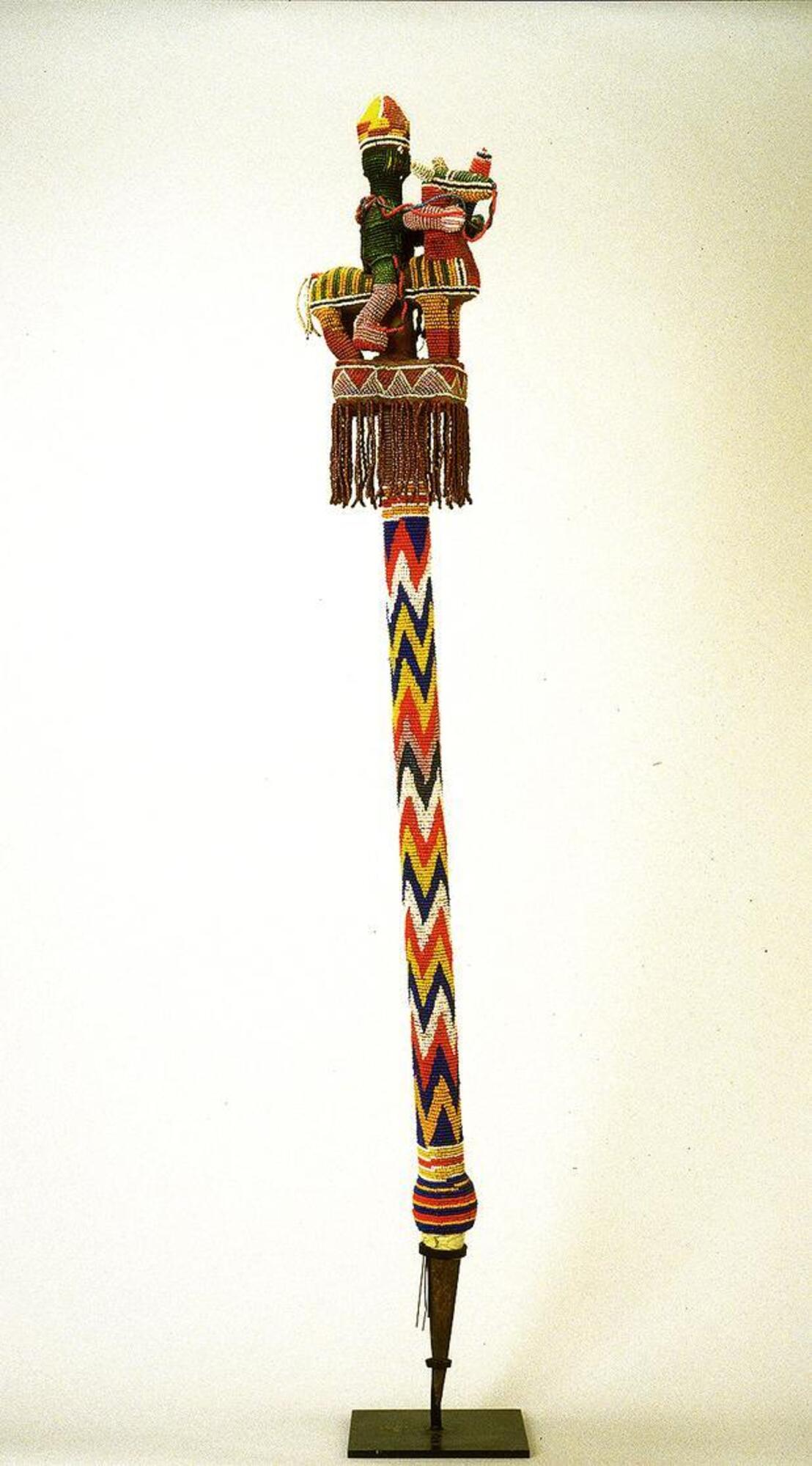 Wooden staff covered with beads, displaying a zig-zag pattern in blue, white, red, yellow, and pink along the shaft, with a metal point at the base. The finial (top of the staff) consists of an equestrian figure, mostly in green, wearing an elaborate red-and-yellow crown, holding a blue staff and riding a multicolored horse, which stands on a rectangular platform adorned with a fringe of beads.