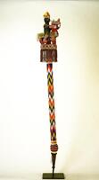 Wooden staff covered with beads, displaying a zig-zag pattern in blue, white, red, yellow, and pink along the shaft, with a metal point at the base. The finial (top of the staff) consists of an equestrian figure, mostly in green, wearing an elaborate red-and-yellow crown, holding a blue staff and riding a multicolored horse, which stands on a rectangular platform adorned with a fringe of beads.