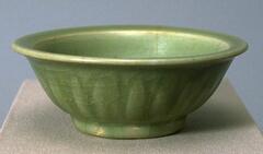 A small stoneware bowl on a footring with an everted, flat rim. It is carved on the exterior to resemble lotus petals, and covered in a green celadon glaze. 