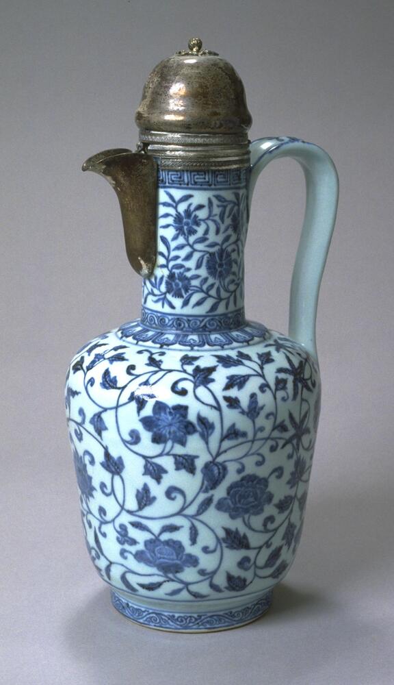 A porcelain ewer with a cylindrical body on a foot ring and a tall, narrow neck. It has a flat, wide, ear-shaped handle, covered with underglaze blue floral meander and clear glaze, and a replacement silver spout and domed lid.