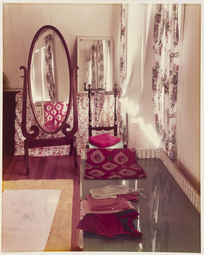 Photograph of an interior view of a room. On the right side of the image is a wicker dresser with folded clothing and a full length wooden oval mirror standing next to the far wall. The window on the right has a long floral curtain.