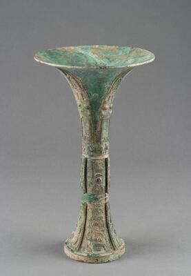 Wine drinking goblet or beaker with a wide, trumpet-shaped mouth, narrow, banded waist, and flaring foot. The slender silhouette of the vessel suggests a date towards the end of the Late Shang period. The body is decorated with Tao-tie mask design, divided by the elaborate raised flanges. An inscription is found inside the flaring foot, presumably the name of the person that the vessel is dedicated.