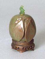 A round jasper snuff bottle with carved decorations of leaves in raised relief. On top of the bottle is a green glass stopper carved in the shape of a bird.