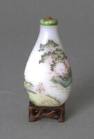 A opaque milk glass snuff bottle in the shape of a teardrop. Decorated on the surface of the snuff bottle is a landscape scene with trees, people, and body of water.