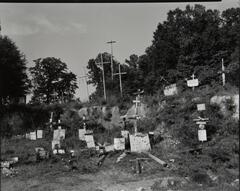 Yard of signs and crosses, hill behind seems to be made of rock, trees above.
