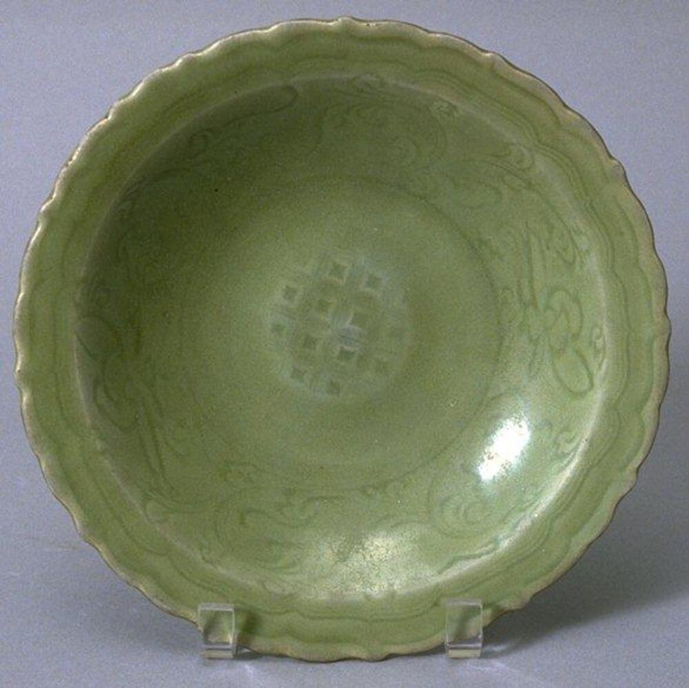A stoneware, shallow bowl with flattened base on a footring with molded sixteen panel foliate rim. The interior is incised with floral meander and a central roundel with a diaper and dot pattern. The bowl is covered in a green celadon glaze.