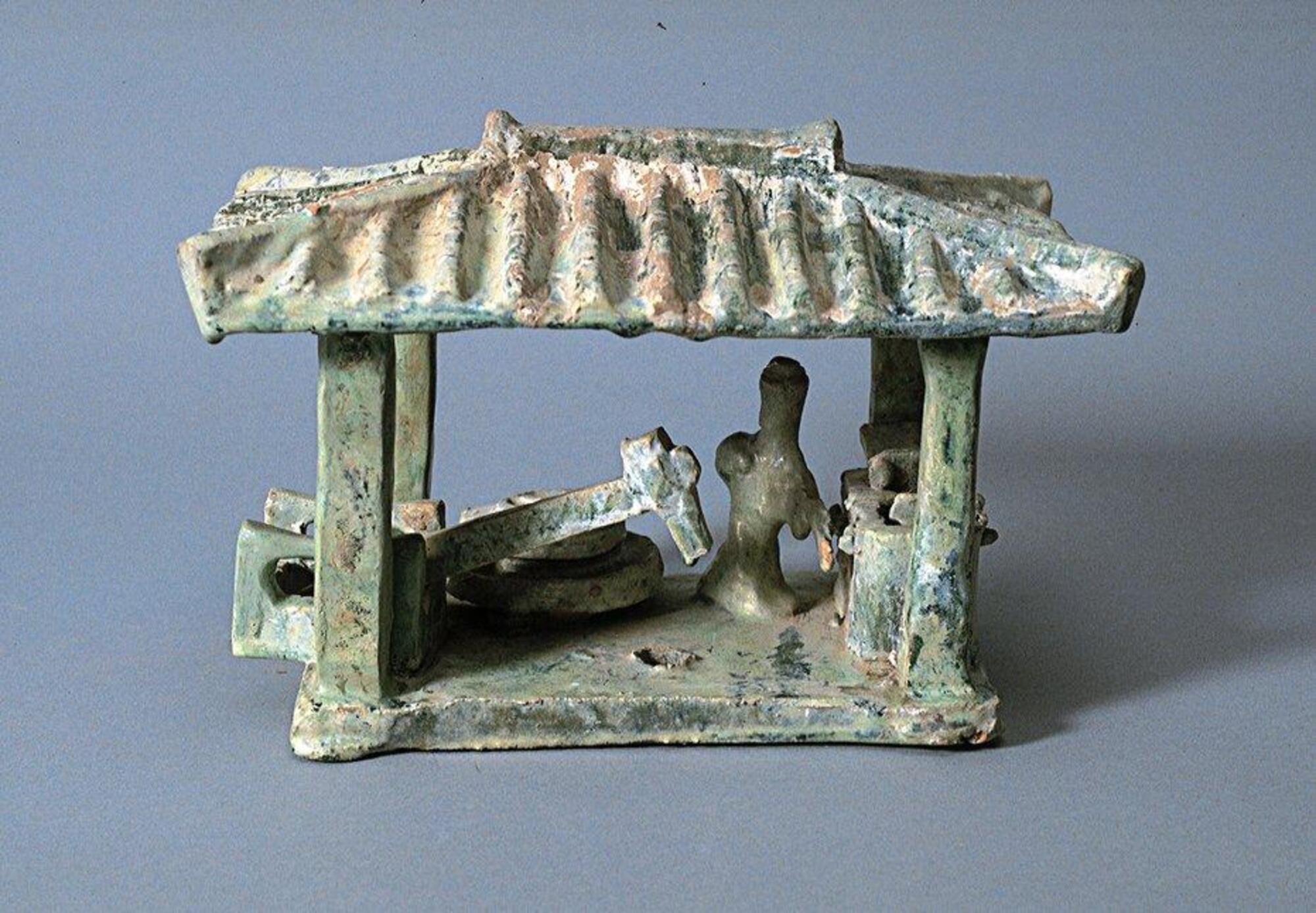 This model of a hulling mill consists of a treadle operated tilted pole connecting to a hammer that crushes the grain, with an operator and storage bins. It sits below a four-posted one-bay structure with a hipped roof with ridge lines, imitating contemporary tiled roofs. The model is covered in a green lead glaze with iridescence and calcification.