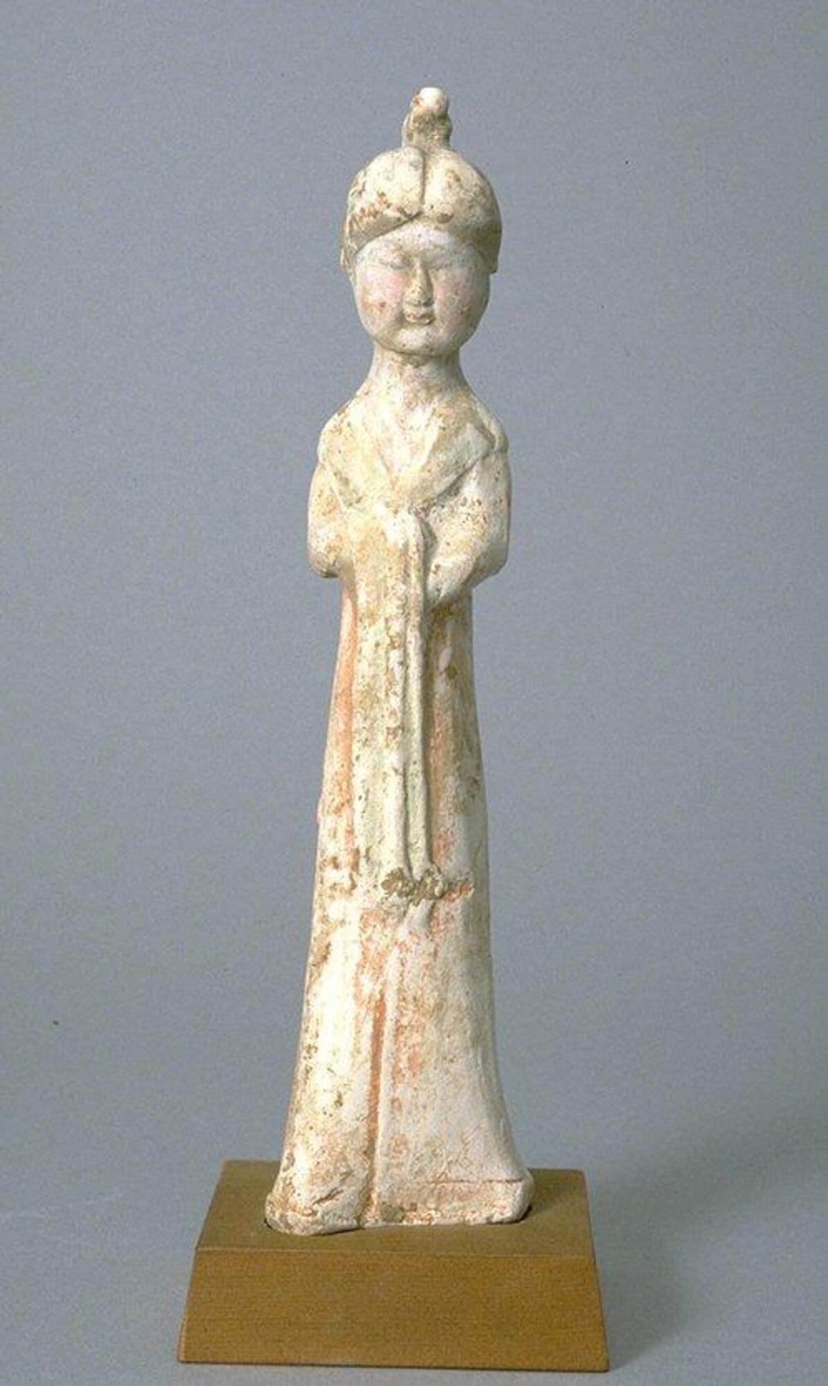 An earthenware female figure, thin and tall, standing with arms clasped in front of the body, wearing long robes with a wide collar, tied at the waist, with the fabric of her long sleeves folded over her hands. She has a round face with petite details, her hair is parted down the middle and tied up in a high chignon. The sculpture is covered in polychrome mineral pigments.