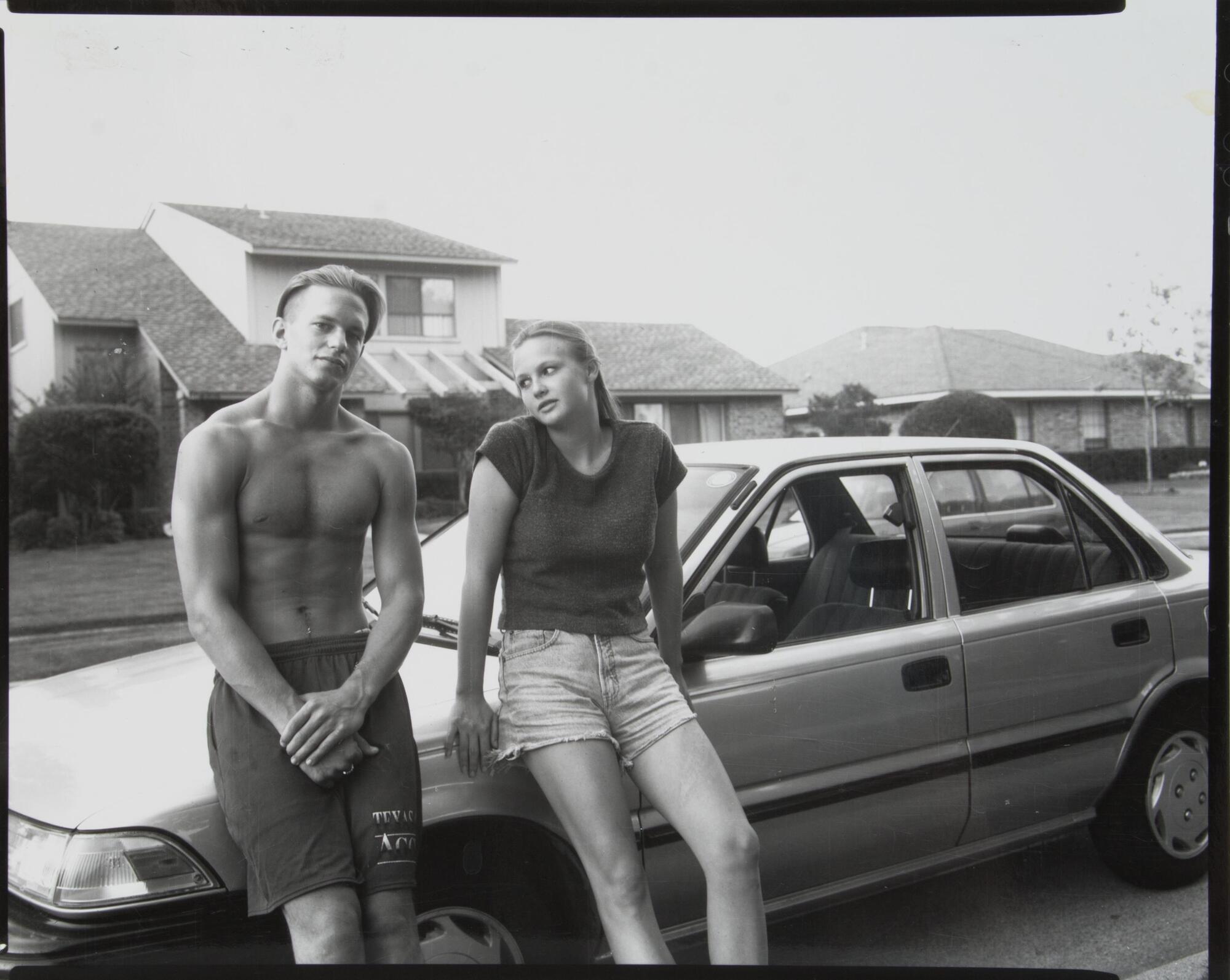 A boy and girl up against a car, girl looking at boy, boy looking straight ahead.