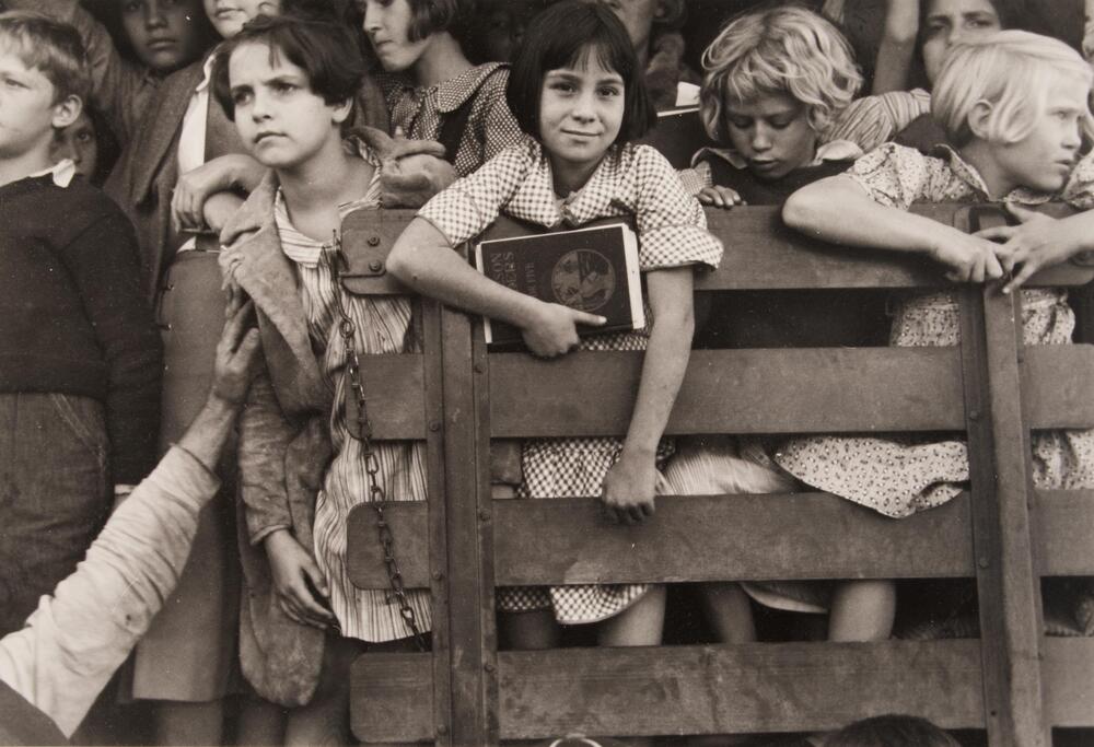 A group of children stand tightly packed together behind a wooden fence.  None of the children appear to be facing the camera except for one girl in a gingham dress at the center of the photo.  This girl, slightly smiling, displays the cover of a book she holds in her hand.