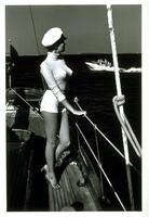 This photograph depicts a woman standing on a sailboat deck, leaning on the railing and looking out at the water as another boat passes by in the background.