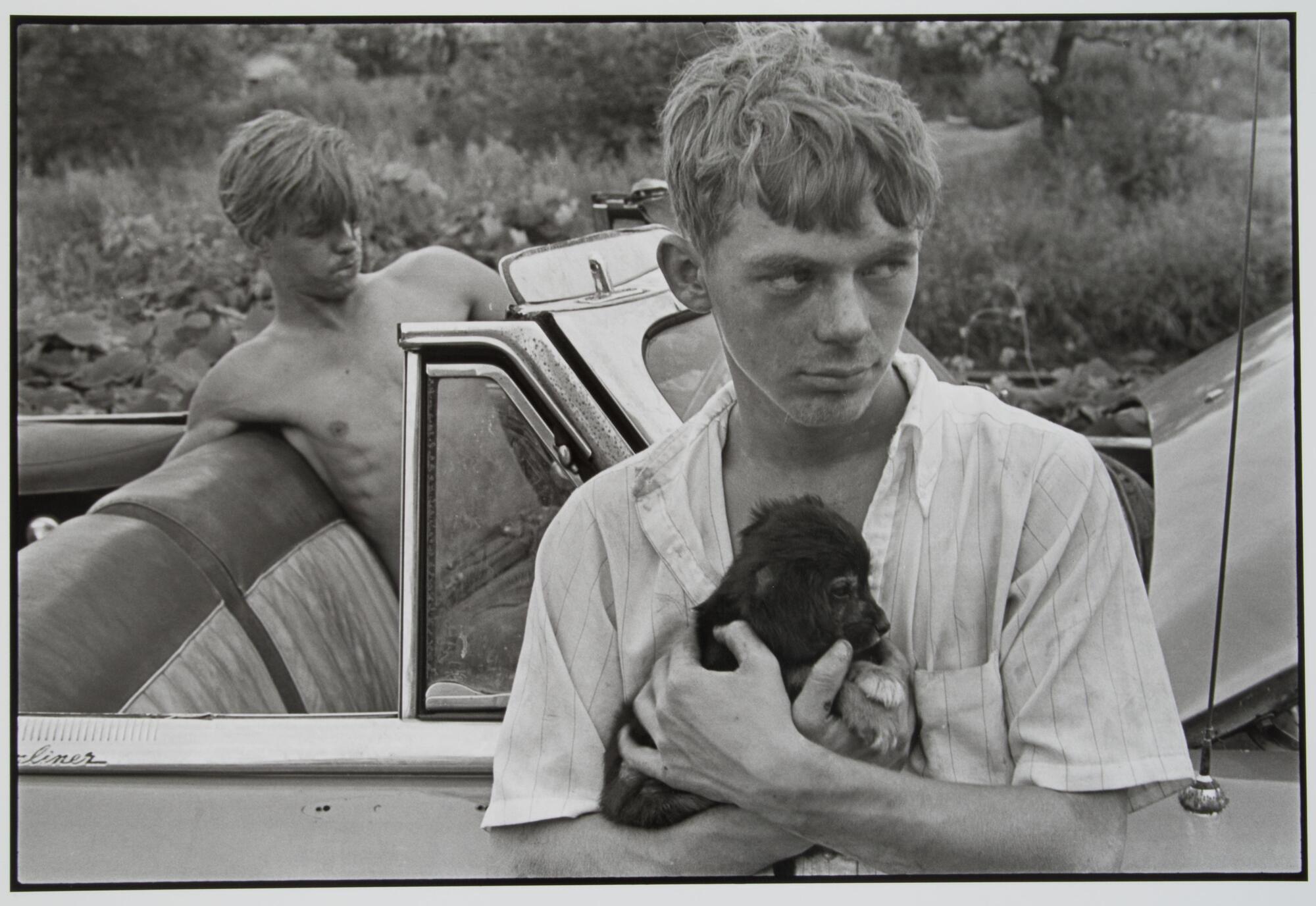 Photograph of a boy holding a puppy. He leans against a car, in which another boy sits in the driver seat, leaning back against the seat.