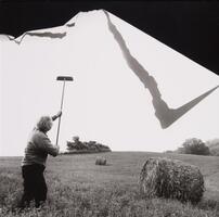 Woman holding a paint roller on a stick in the air in the middle of a field, top of the photograph looks like it is folding down.