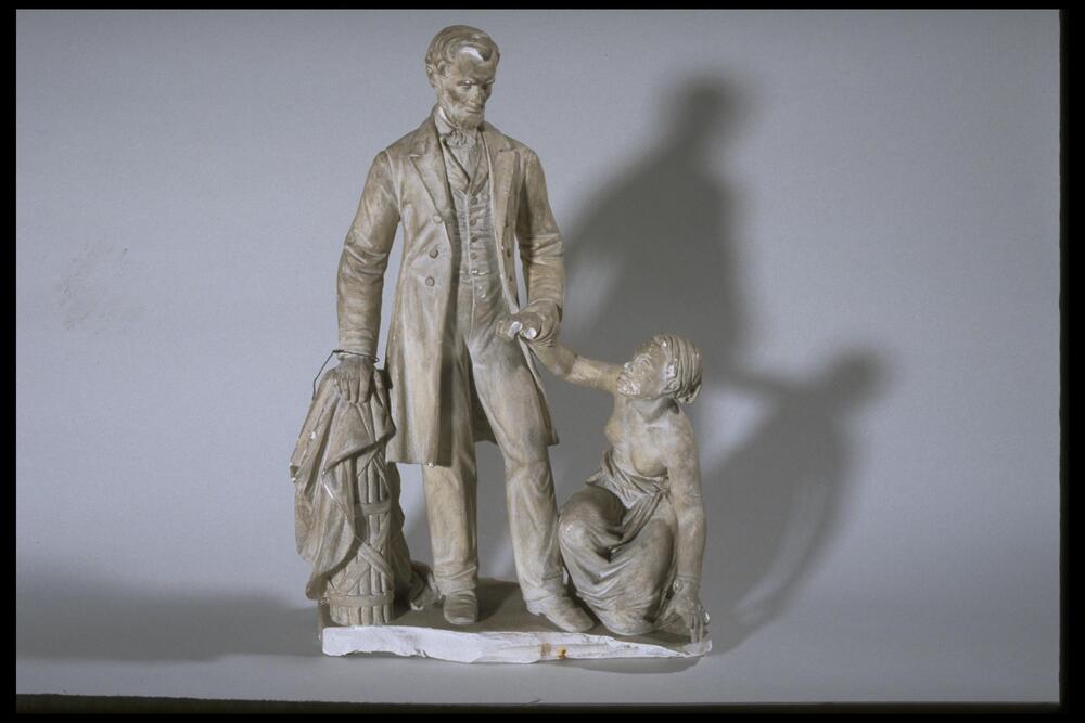 Plaster sculpture of a standing male figure, his right hand resting upon a small column and his left hand holding the arm of a crouching African American female figure.