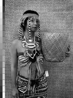 Black and white photograph of a woman holding a woven basket. She stands in front of a spotted background and wears a striped bandeau, a wrapped skirt with a geometric design and the price tag still attached, a necklace strung with large ornaments, and headdress with cloth strips or beads that hang over her face and the back of her head.&nbsp;