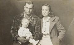 A family portrait of a man, woman, and infant. The man and woman stand, the man on the left is wearing a plaid jacket, and the woman on the right is in a coat, blouse, and skirt. The infant sits in the man's lap. 