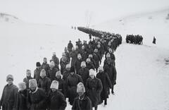 A column of men in long coats and fur hats march through a snow-covered landscape.
