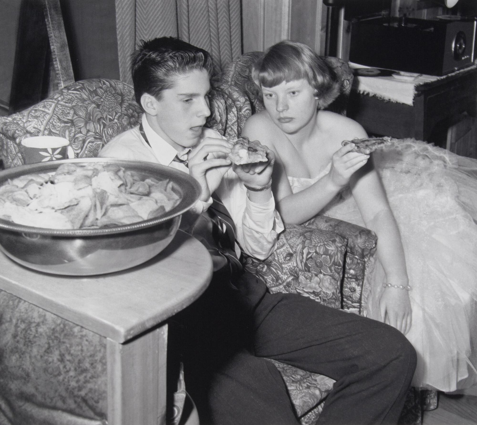 A boy and girl seated on two upholstered chairs eating pizza. The girl is wearing a fancy dress and the boy is in a shirt and tie. There is a large bowl of potato chips on the table.