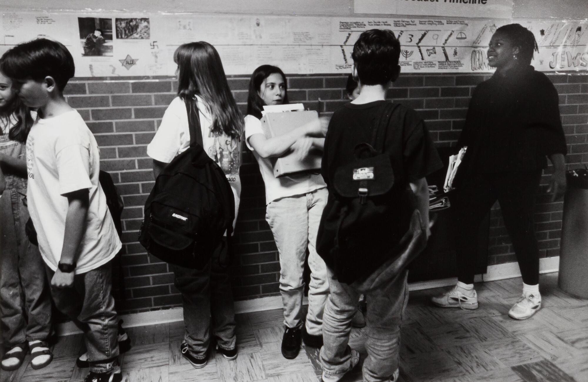 Hallway of a school with children. One girl is leaning on a wall holding large binders, posters on the wall.