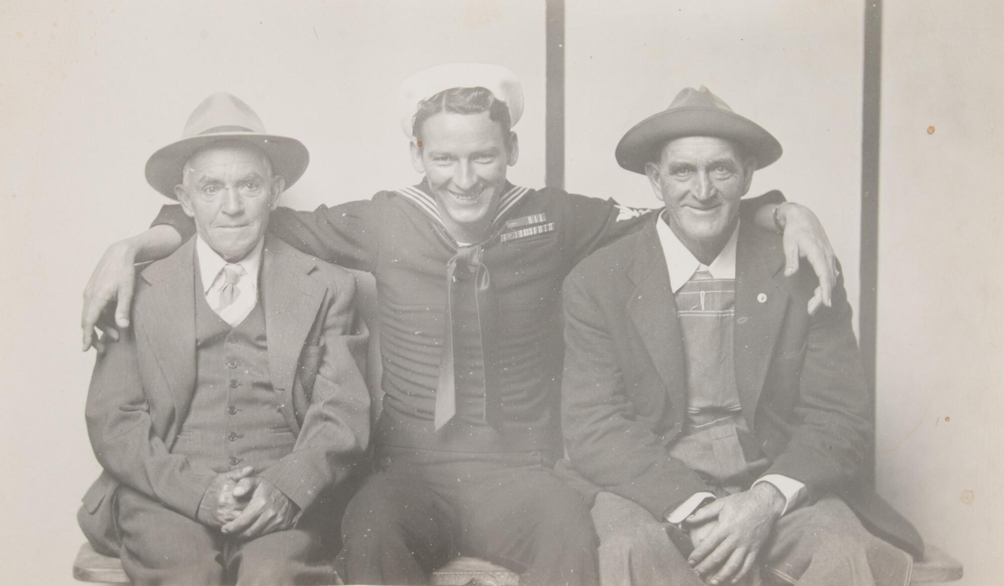 A portrait of three seated men. On the left, an older man sits with his hands clasped in his lap. In the center, a man in naval uniform sits with his arms around both men. On the right, a third man sits with his hands in his lap. All three wear hats and smile.