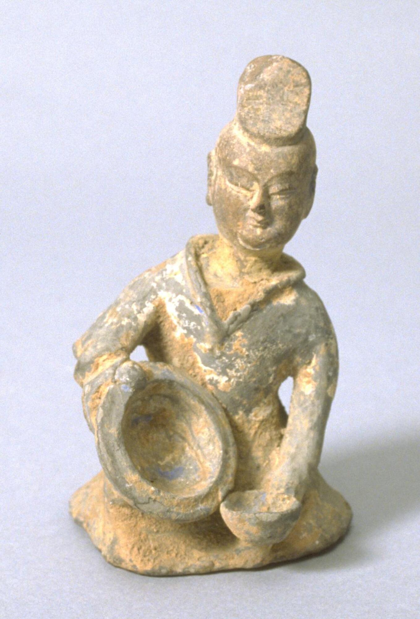 This is s clay figure of a seated woman wearing an officials cap and a robe. She is holding two bowls, shown in the action of pouring the contents from the larger bowl into the smaller bowl.