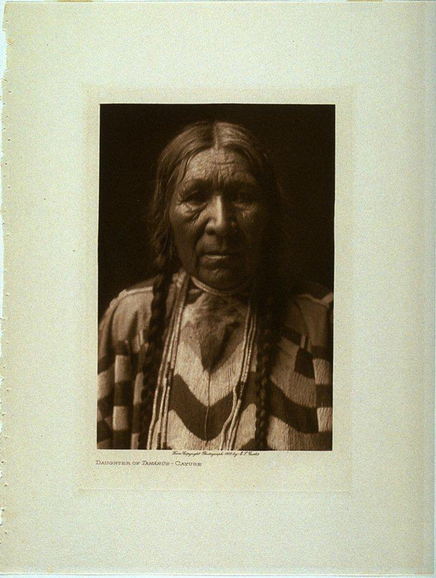 A portrait of an aging woman. She looks directly into the lens, wearing braids, strands of beaded necklaces, and a garment with geometric lines.