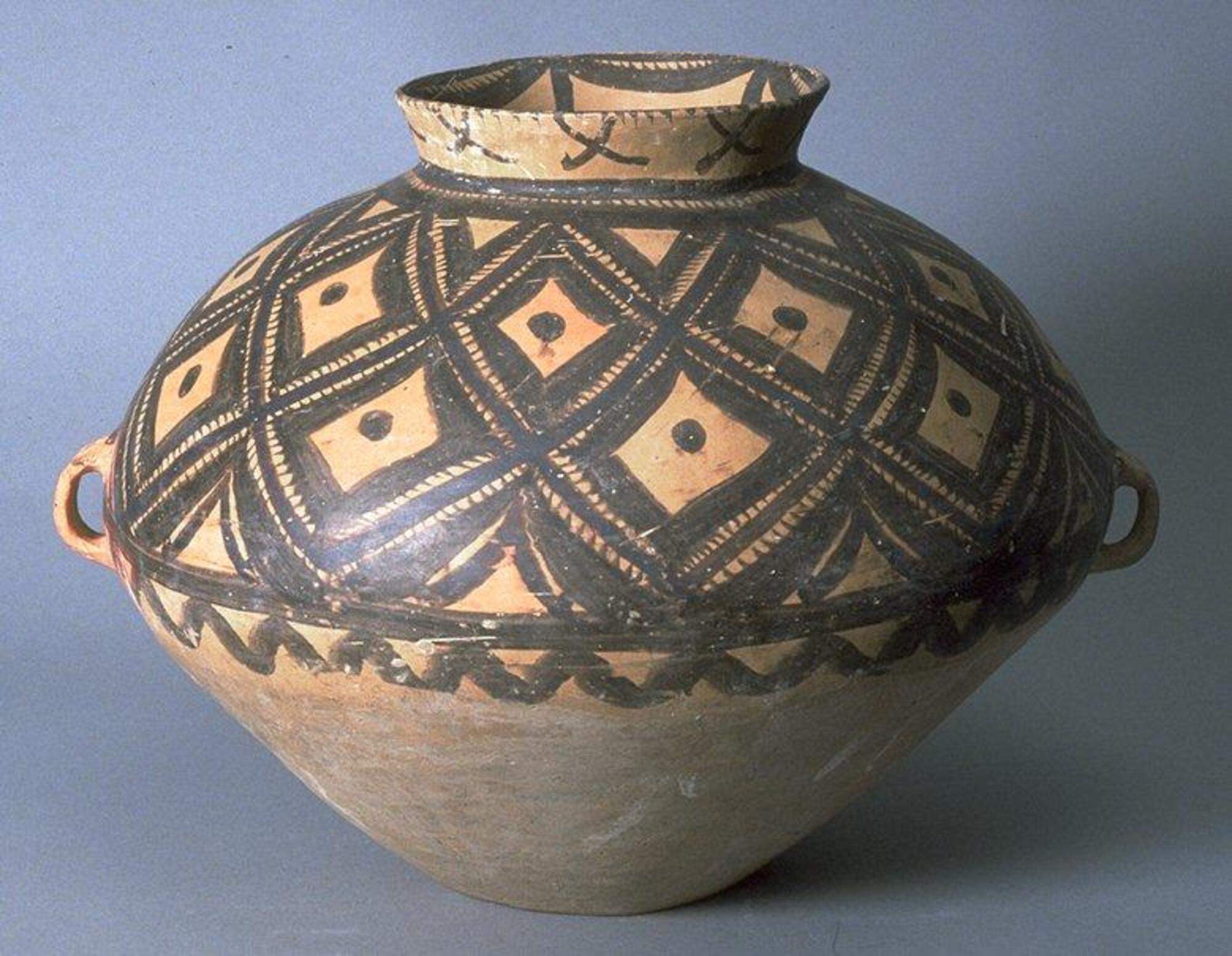 This light reddish-buff earthenware <em>guan (</em>罐) jar has a wide globular upper body and a conical lower body on a narrow flat base with a narrow neck and flaring rim. There are two diametrically opposed lug handles at the waist. Painted on the upper half of the body with black pigment are a network of rhomboid shapes, hatch-marks and dots confined within solid band borders, with a lobed line border below.  Around the neck are a painted "X" design, hatch marks and a lobed border to the interior.  